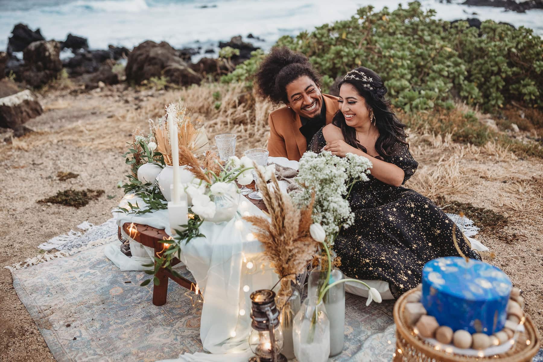 Wedding & Elopement Photographer, man and woman sit at festive table by the sea, it is decorated with floral bouquets and food