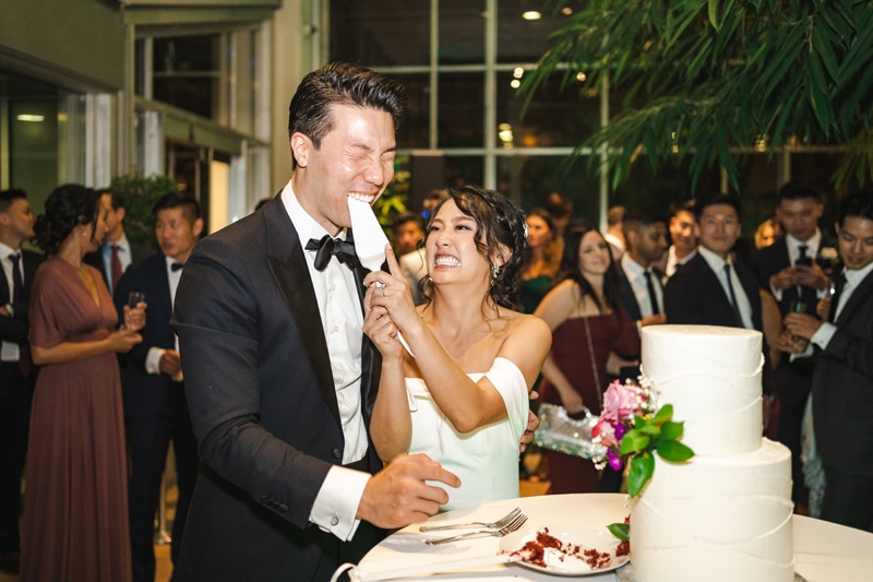 Wedding and Elopement Photography, bride feeds groom wedding cake with cake server in fun, crowd watches